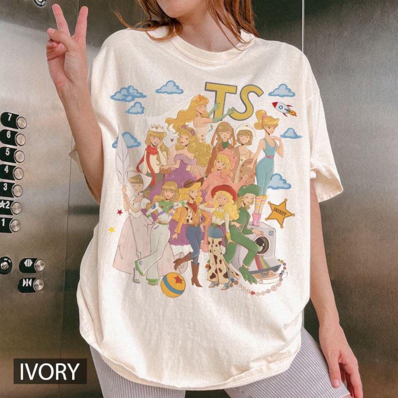 K&ouml;nnte beinhalten: Ein elfenbeinfarbenes T-Shirt mit einem Grafikdruck verschiedener animierter Figuren im Collage-Stil. Das Design enth&auml;lt die Buchstaben TS, eine Rakete, einen Sheriff-Stern und einen bunten Ball. Die Figuren tragen verschiedene Outfits.