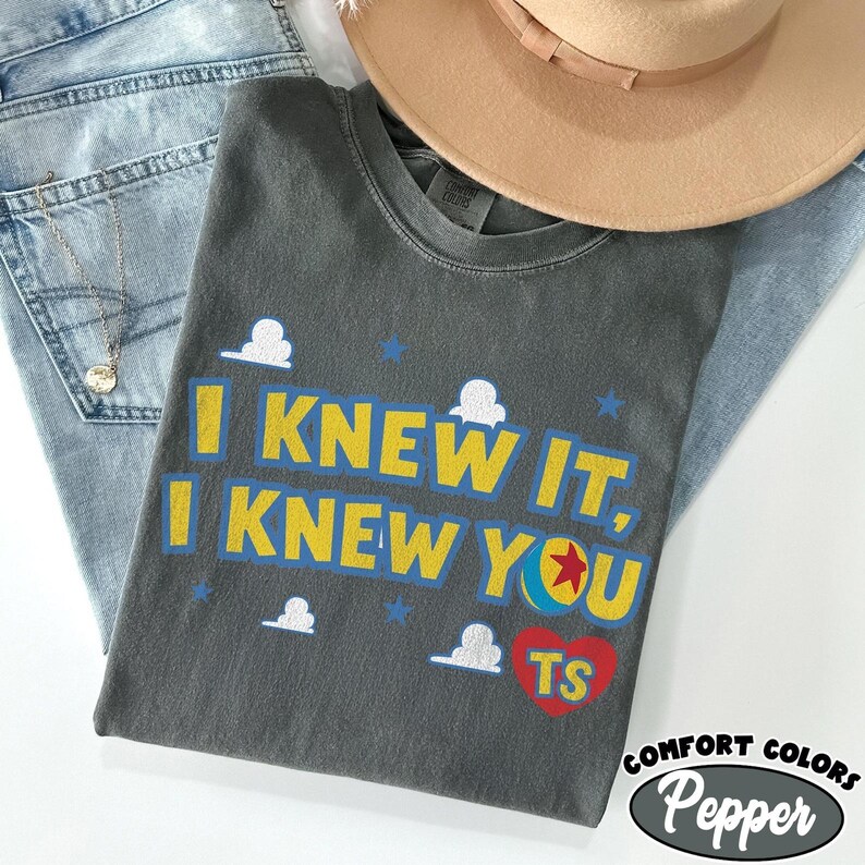 May include: A charcoal gray short-sleeve t-shirt featuring the yellow text I KNEW IT, I KNEW YOU with a blue outline. The design includes white cloud graphics, small blue stars, a colorful ball with a red star, and a red heart containing the letters TS.
