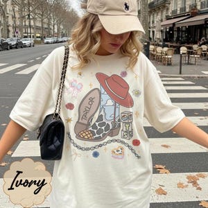 May include: An ivory t-shirt featuring a graphic print of a cowboy boot with a cow print pattern, a red hat, a bow, and blocks with the letters T, S, and 13. The boot has the name Taylor printed on the sole. A black shoulder bag is also visible.