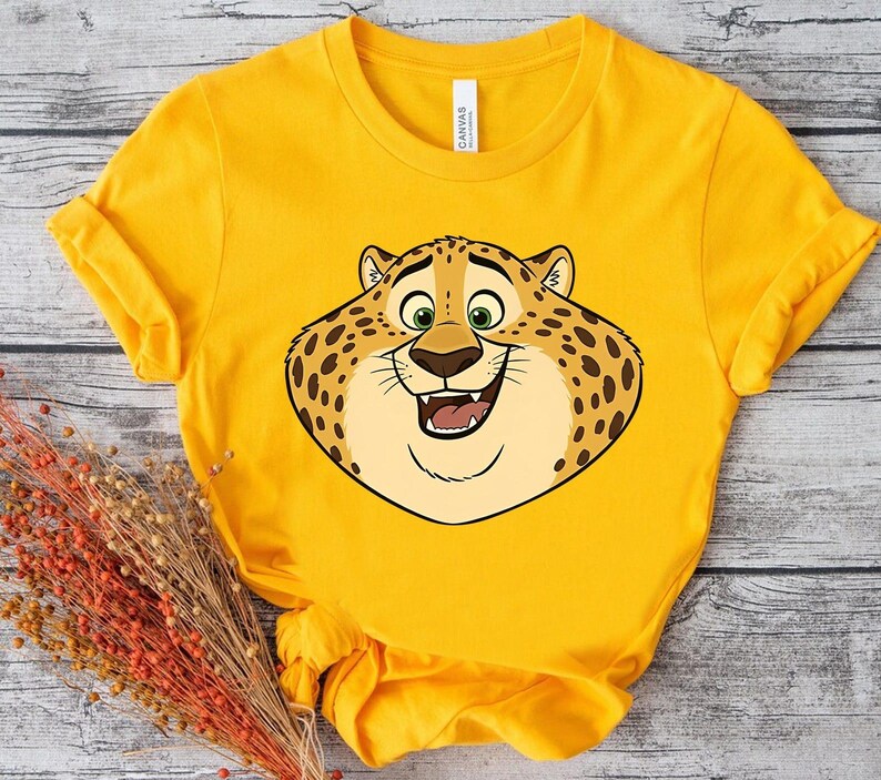 K&ouml;nnte beinhalten: Ein gelbes T-Shirt mit einem Cartoon-Leoparden-Gesicht, das l&auml;chelt. Der Leopard hat braune Flecken, gr&uuml;ne Augen und eine hellbraune Nase. Das T-Shirt besteht aus einem weichen Material.