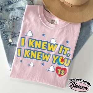 May include: A light pink short-sleeved t-shirt featuring the blue and yellow text I KNEW IT, I KNEW YOU with a red heart containing the letters TS, a colorful ball graphic, and small white cloud and blue star icons printed on the front chest area.