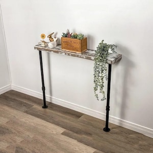 May include: Whitewashed wooden console table with black metal pipe legs. The table is decorated with a small wooden box with the text "PSALM 16:11" and a small succulent plant. A small white rabbit figurine and a yellow flower are also on the table. A trailing green plant hangs from the table.