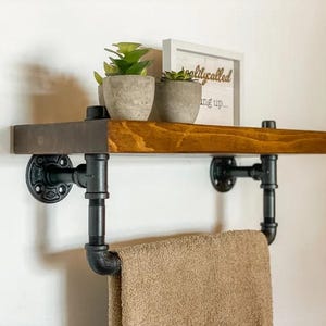 May include: A wooden shelf with black metal pipe brackets and a towel rail. The shelf is mounted to a white wall and has two small potted plants and a sign that reads "reality called..." on it.