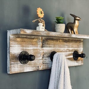 Könnte beinhalten: Rustikales Holzregal mit drei schwarzen Haken, einer goldenen Hasenfigur und einer dekorativen Blume. Das Regal besteht aus Altholz mit weißen Farbakzenten. Ein weißes Handtuch hängt an einem der Haken. Ca. 60 cm breit.