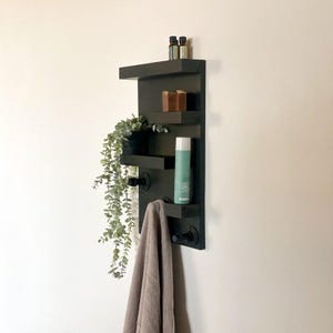Peut inclure: Étagère murale en bois noir avec trois étagères et deux crochets. L'étagère a une serviette accrochée à un crochet et une plante sur l'étagère supérieure. Il y a aussi des bouteilles et du savon sur les étagères.