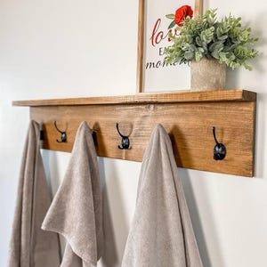 May include: A wooden shelf with five black hooks, holding beige towels. The shelf is 36 inches wide and Mission Oak in color. Above the shelf is a framed print with a red rose and the words "love each moment".