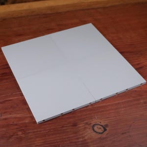 May include: A light grey, square panel, possibly a tile, is shown. It's divided into four sections by subtle lines. The edge has small, evenly spaced holes. The item rests on a dark wooden surface. The panel's dimensions are approximately 30 cm x 30 cm.