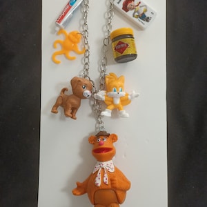 Peut inclure: Un collier à chaîne en métal argenté orné de plusieurs breloques miniatures, dont un tube de dentifrice Colgate, une pieuvre jaune, un pot de Vegemite, un domino Jessie de Toy Story, un chiot marron, une figurine Tails jaune et Fozzie Bear.