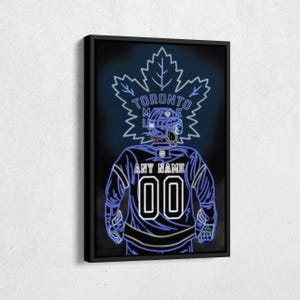 May include: A black framed canvas print with a neon blue design of a hockey player wearing a Toronto Maple Leafs jersey. The player's back is facing the viewer and the jersey has the text "ANY NAME" and the number "00" on the back. The design also includes a neon blue maple leaf with the text "TORONTO MAPLE LEAFS" around it.