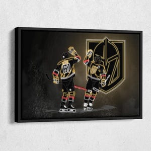 May include: A framed art print featuring two hockey players in black and gold uniforms, celebrating with a high-five. The background is a gradient of dark gray and gold, with the team logo prominently displayed. The artwork has a neon-like effect.