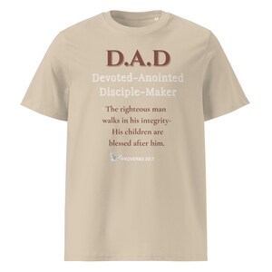 May include: A beige short-sleeved t-shirt featuring the brown text D.A.D, white text Devoted-Anointed Disciple-Maker, and the verse The righteous man walks in his integrity- His children are blessed after him. Proverbs 20:7 with a small dove icon.
