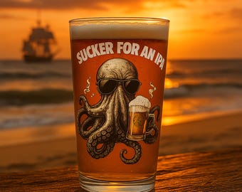 May include: A pint glass filled with beer, featuring an octopus wearing sunglasses and holding a beer mug. The text "SUCKER FOR AN IPA" is above the octopus. The background shows a beach scene with a ship at sunset.