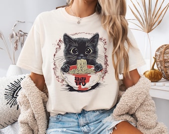 May include: A white t-shirt with a black cat eating ramen noodles from a red bowl. The cat has big eyes and is wearing a red collar. The bowl has Japanese characters on it.