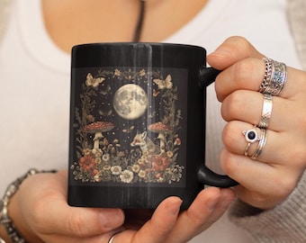 May include: Black ceramic mug with a forest scene design. The design includes a full moon, mushrooms, butterflies, and a fox in shades of brown, beige, and red. The mug has a black handle and a glossy finish.