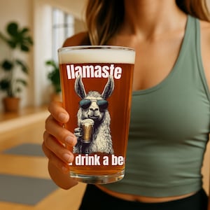 Op de afbeelding: Een helder glazen bierglas met een lama die een zonnebril draagt en een biertje vasthoudt. Het glas heeft de tekst "Llama-ste drink a beer" erop gedrukt.
