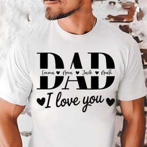 Puede incluir: Una camiseta blanca de manga corta con el texto negro DAD en una fuente grande y negrita. Centrados dentro de las letras están los nombres Emma, Nova, Jack y Noah separados por corazones. Debajo, el texto I love you flanqueado por dos corazones.