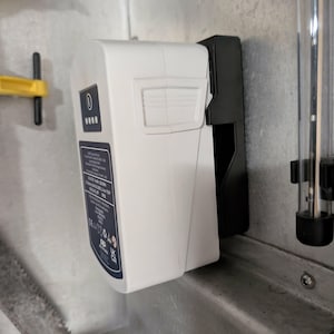 May include: A white rectangular device with a black mounting bracket, attached to a metal surface. The device has a small display and a label with text. A yellow handle and a clear tube are also visible.