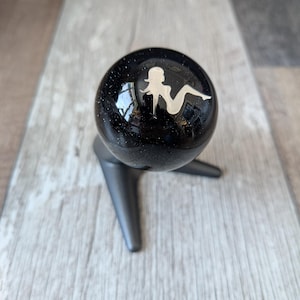 May include: A black sphere with a silver silhouette of a woman, mounted on a black three-legged stand. The sphere has a speckled texture and reflects light. The stand is a matte black finish. The object is on a wooden surface.