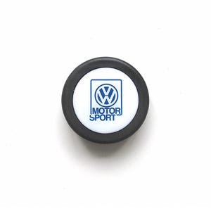 May include: A round, black and white emblem with the words "MOTOR SPORT" in blue text. The emblem features a blue Volkswagen logo in the center.