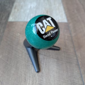 Peut inclure: Un pommeau de levier de vitesses scintillant vert d'eau avec le logo CAT Diesel Power. Le pommeau est rond et repose sur une base noire à trois pieds. Le logo est noir et blanc avec un triangle jaune.