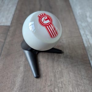 May include: A white spherical gear shift knob featuring a red Cummins logo with vertical stripes. The knob is mounted on a black, three-pronged tripod stand. The design includes the word Cummins printed in red within the circular logo emblem.