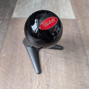 Peut inclure: Un pommeau de levier de vitesses sphérique noir avec le logo "Peterbilt" rouge et blanc. Le bouton est monté sur une base noire à trois branches. Le fond est une surface en bois brun clair.
