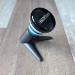 May include: A black gear shift knob featuring the word FREIGHTLINER in white text on the top. The knob has a glossy finish with horizontal blue and white stripes below the top section and is mounted on a matte black three-pronged metal stand.
