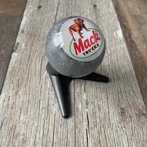 May include: A silver glitter ball with the Mack Trucks logo, featuring a bulldog illustration, set on a black stand. The logo is in a white circle with a green border, and the text "Mack TRUCKS" in red.