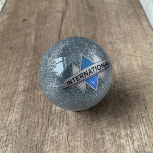 Peut inclure: Une sphère grise scintillante avec le mot "INTERNATIONAL" imprimé sur un logo bleu et blanc. La sphère est posée sur un fond en bois.