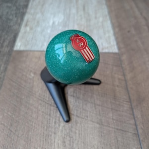 Peut inclure: Une sphère scintillante turquoise avec un emblème rouge et blanc, montée sur un support noir à trois pieds. L'emblème présente les lettres "KW" et un design stylisé. La sphère est le point central, avec une finition brillante.