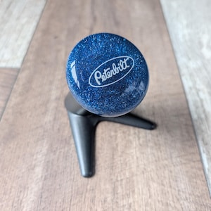 Peut inclure: Un pommeau de levier de vitesse rond bleu foncé avec une finition pailletée et le logo blanc Peterbilt imprimé sur la surface supérieure. Le pommeau est fixé sur une base métallique noire à trois branches qui soutient l'élément sphérique.