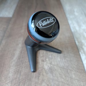 Peut inclure: Pommeau de levier de vitesse Peterbilt noir et argenté avec le logo Peterbilt en blanc. Le pommeau a un dessus arrondi avec une bande argentée et rouge. La base est de couleur gris foncé.
