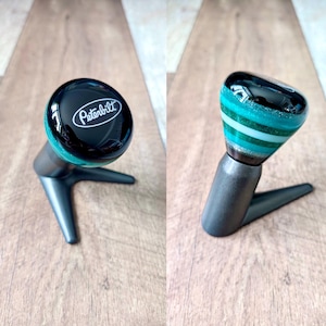 Peut inclure: Un pommeau de levier de vitesse Peterbilt personnalisé avec un dessus noir et un design à rayures turquoise et blanches. Le pommeau est monté sur une base en métal gris foncé avec un design à trois branches.