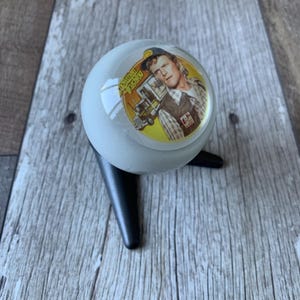 May include: A decorative, round, light gray object with a black stand. The object features a circular image of a man in a trucker hat and the words "Snowman Trucking". The image is set against a yellow background.