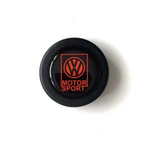 Puede incluir: Un emblema negro y redondo con el logotipo rojo de Volkswagen dentro de un cuadrado rojo y las palabras "MOTOR SPORT" en blanco. Probablemente un accesorio para el coche.