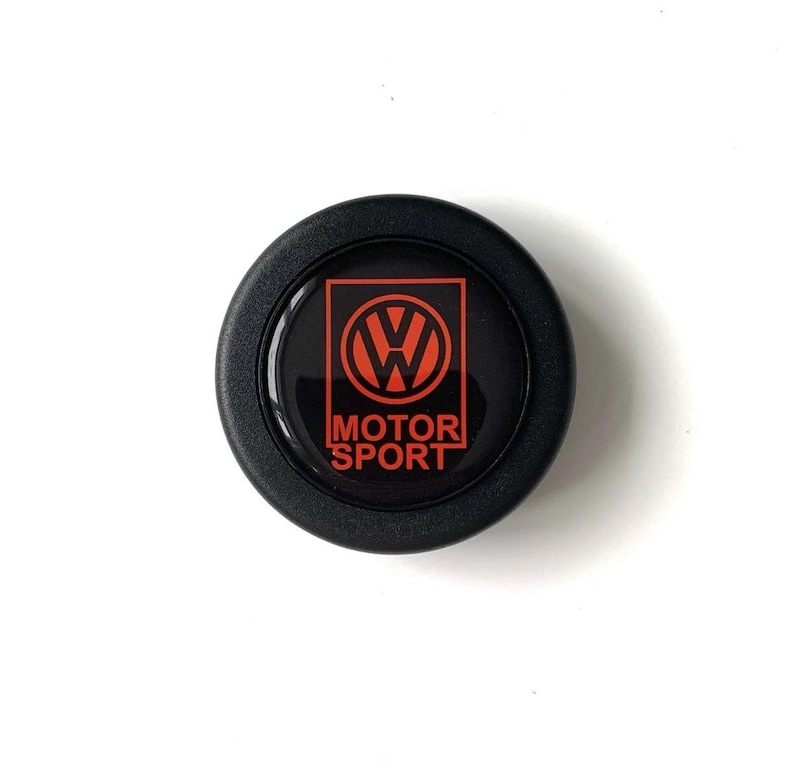 Puede incluir: Un emblema negro y redondo con el logotipo rojo de Volkswagen dentro de un cuadrado rojo y las palabras "MOTOR SPORT" en blanco. Probablemente un accesorio para el coche.
