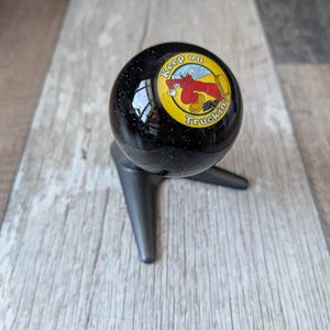 May include: A black, speckled shift knob with the text "Keep on Truckin'". The graphic features a cartoon character in red and yellow. The shift knob is mounted on a dark grey, three-legged stand.