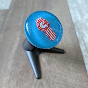 May include: A blue gear shift knob with a glittery finish and a red and white Cummins logo on the top. The knob is attached to a black, three-pronged base. The circular top features the red Cummins text and emblem with vertical stripes.