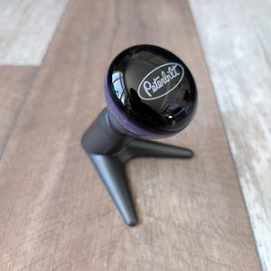 May include: A black Peterbilt gear shift knob with a purple accent ring. The knob has a glossy finish and the Peterbilt logo in white. The base is a dark gray, three-pronged design.