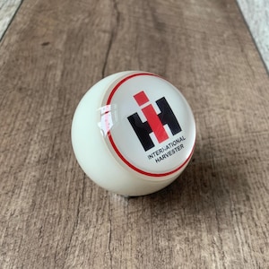 Peut inclure: Un pommeau de levier de vitesses International Harvester blanc et rond. Le pommeau présente le logo IH en noir et rouge, avec les mots "INTERNATIONAL HARVESTER" en noir. Un anneau rouge entoure le logo. Le pommeau est posé sur un fond en bois.