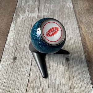 Peut inclure: Un pommeau de levier de vitesses bleu sarcelle scintillant avec le logo Peterbilt dans un cercle blanc bordé de rouge. Le pommeau est monté sur une base noire. Le logo est centré sur le dessus.