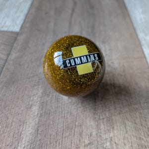May include: A spherical gear shift knob featuring a gold glitter finish throughout the surface. A rectangular logo is embedded in the centre, displaying the word CUMMINS in white capital letters against a black background, set over a yellow bar.