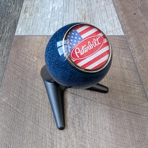 Peut inclure: Un pommeau de levier de vitesses bleu et argenté avec le logo Peterbilt et un drapeau américain. Le pommeau rond a une bordure dorée. La base noire a un design à trois branches.