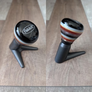May include: A black and multi-colored gear shift knob with the Peterbilt logo. The knob has a black, glossy top and a striped design in shades of brown, white, and gray. It is mounted on a dark gray, angular base.