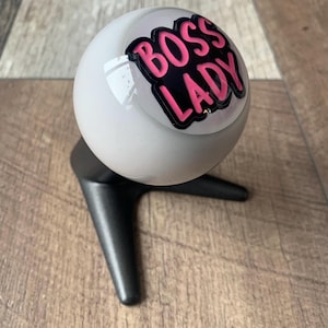 Peut inclure: Un objet sphérique blanc avec le texte "BOSS LADY" en lettres roses soulignées de noir. L'objet repose sur une base noire à trois pieds, sur un fond en bois.