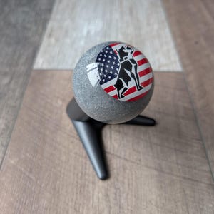 May include: A grey, glittery sphere featuring a black dog silhouette over an American flag design. The sphere is mounted on a black, three-legged stand. The background is a wooden surface.