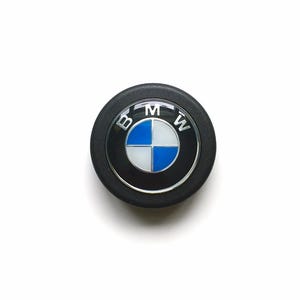 May include: A black and silver BMW emblem with the BMW logo at the centre. The logo features a blue and white quadrant design, surrounded by a silver ring and the letters "BMW".