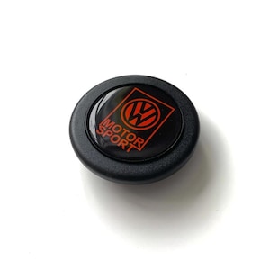 Puede incluir: Un emblema negro y rojo de Motor Sport. El emblema presenta el logotipo de VW en rojo, con las palabras "MOTOR SPORT" en texto rojo. El emblema es circular y tiene un acabado brillante.