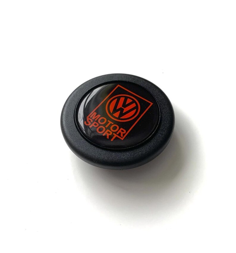 Puede incluir: Un emblema negro y rojo de Motor Sport. El emblema presenta el logotipo de VW en rojo, con las palabras "MOTOR SPORT" en texto rojo. El emblema es circular y tiene un acabado brillante.
