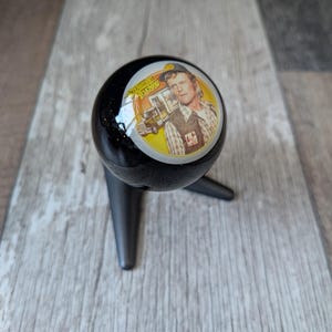 May include: Black gear shift knob with a circular image of a man and a truck. The image is set against a yellow background with the words "Towne's Trucking". The knob is mounted on a black, three-pronged base.